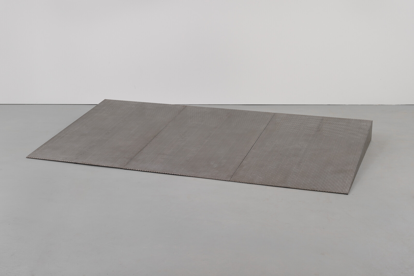 A large rectangular perforated metal sheet lies flat on a bare concrete floor against a white wall.