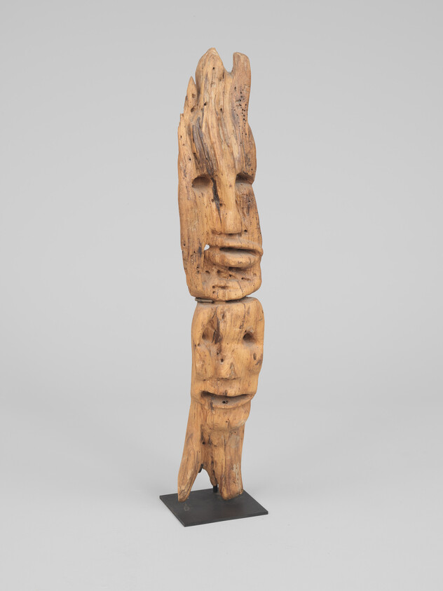 Tall carved wooden sculpture with two stacked abstract human faces on a metal stand.