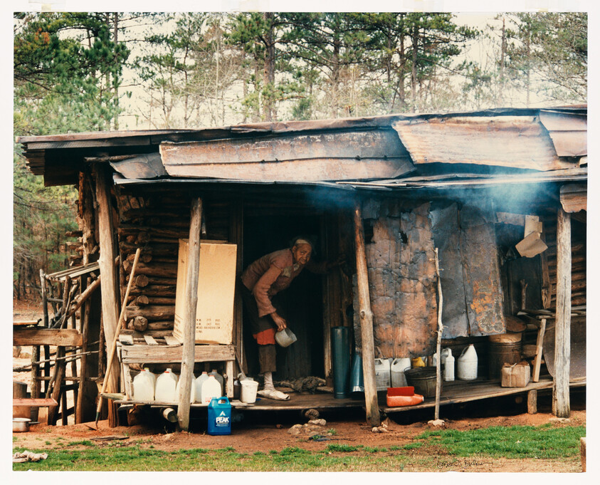An elderly person bends on a rustic cabin porch pouring water from a jug into a container.