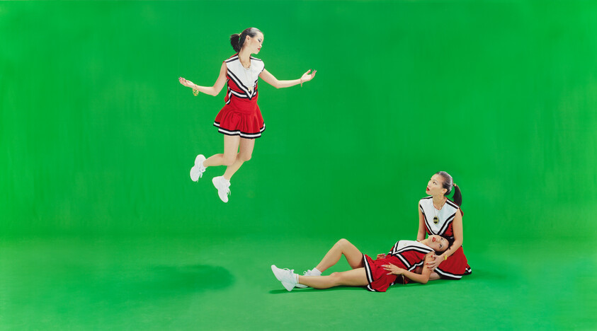 Three cheerleaders in red uniforms on a green background, one jumping while two rest on the floor.