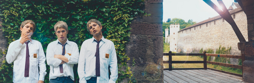 Three teenage students in white shirts and ties stand by an ivy-covered wall, one smoking.