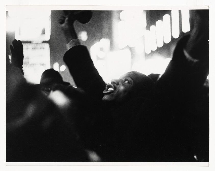 A man in a crowd raises his arms and looks up with an excited expression.