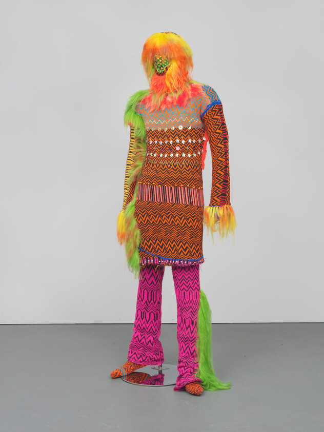 A vibrant and colorful figure covered in textured, patterned fabric and faux fur stands against a white background. The figure's face is obscured by a shaggy, neon orange and green wig, and it wears a long-sleeve, knee-length dress with a zigzag pattern in a multitude of colors. The dress is adorned with fringe at the hem. Matching patterned leggings and shoes complete the ensemble, with one leg covered in long, flowing green fur. The overall effect is a playful and surreal blending of textures and bright colors.