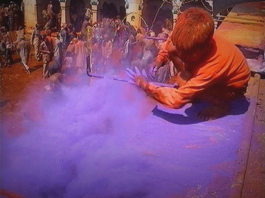 A person pours purple colored powder over a vehicle roof during a crowded street festival.