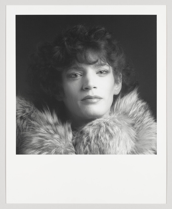 Person with short curly hair wearing a fur collar and looking directly at the camera.