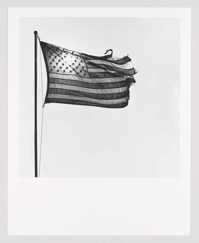 A black and white photograph of an American flag with a slightly tattered edge, flying at full mast against a clear backdrop.