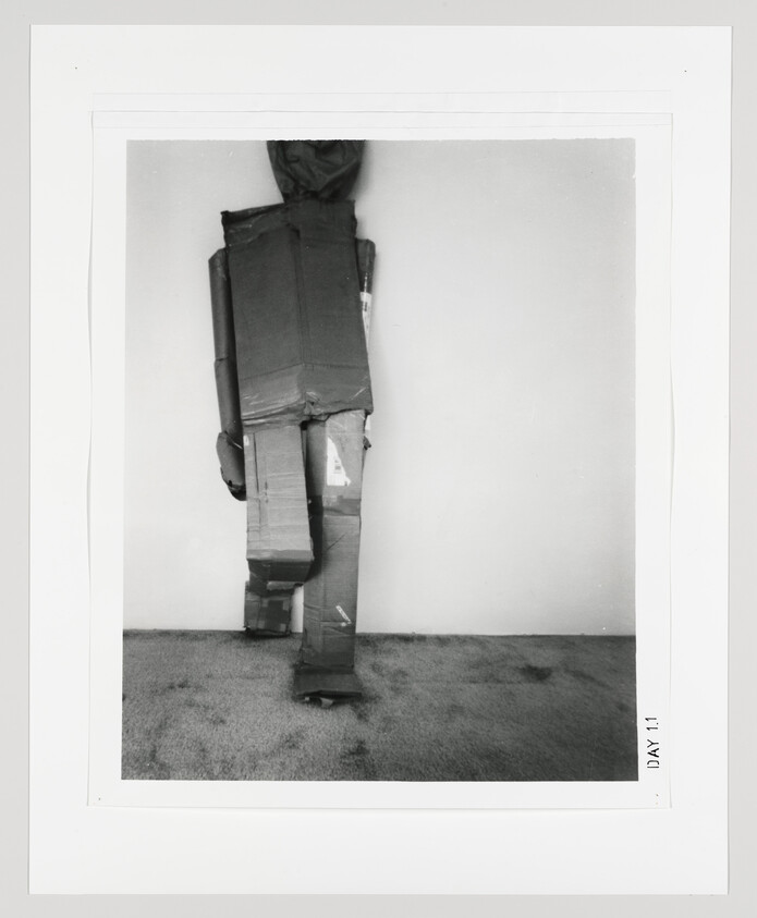 A black and white photograph of a person standing against a wall, covered entirely in cardboard boxes that are arranged to resemble a humanoid figure. The boxes are taped together, and the person's head is also covered with a box, obscuring their identity. The image has a border with the text "DAY 1" printed on the bottom right corner, suggesting it might be part of a series or project. The setting appears to be a room with a carpeted floor.