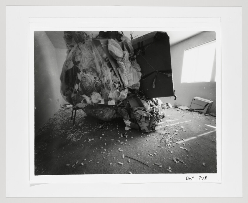 A large compressed pile of trash and debris occupies a sparsely furnished room with light from a window.