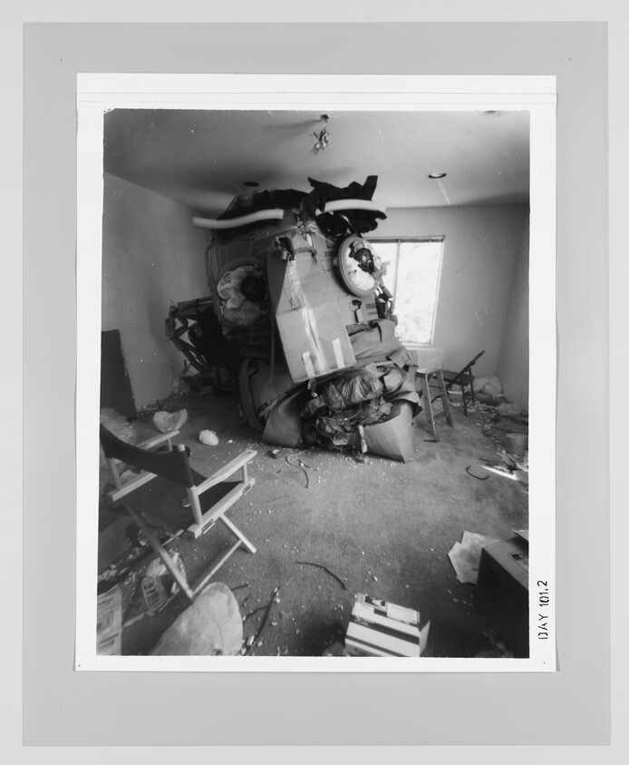 An overturned car crushed into a small living room, scattering furniture and debris across the floor.
