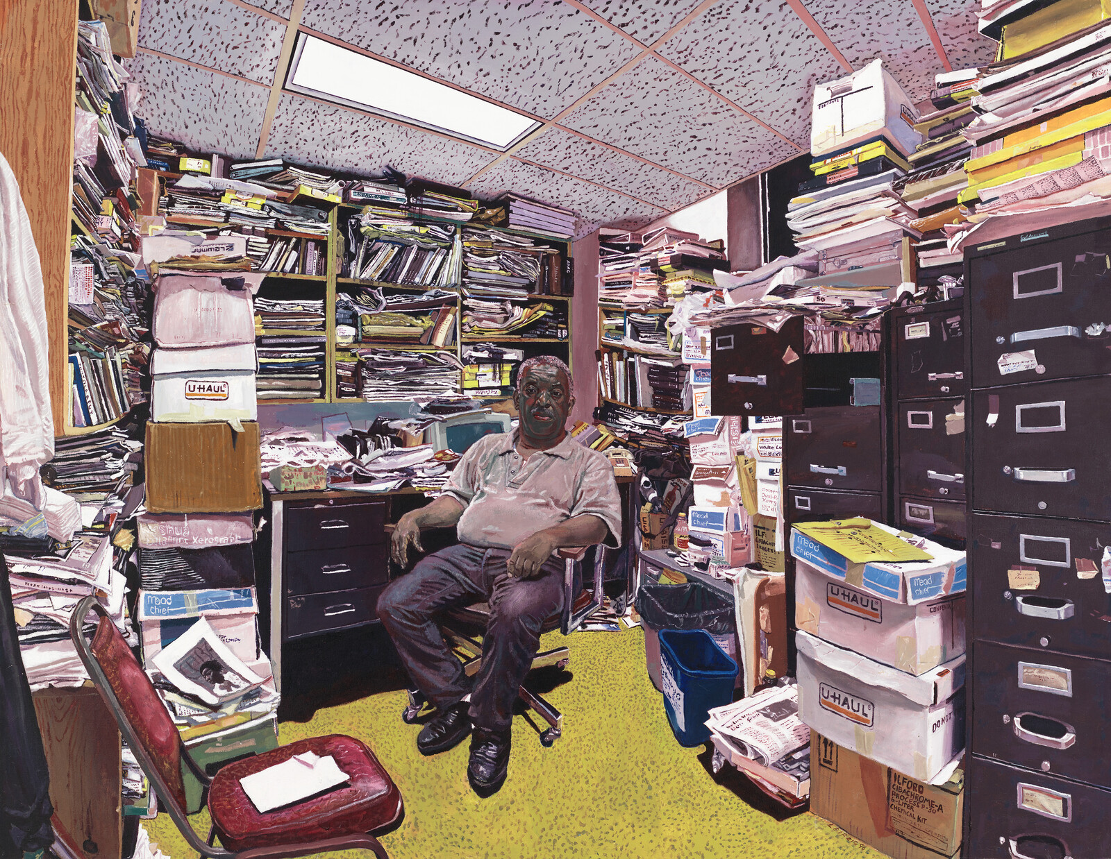 A man sits in a cluttered office surrounded by stacks of papers, filing cabinets, and moving boxes.