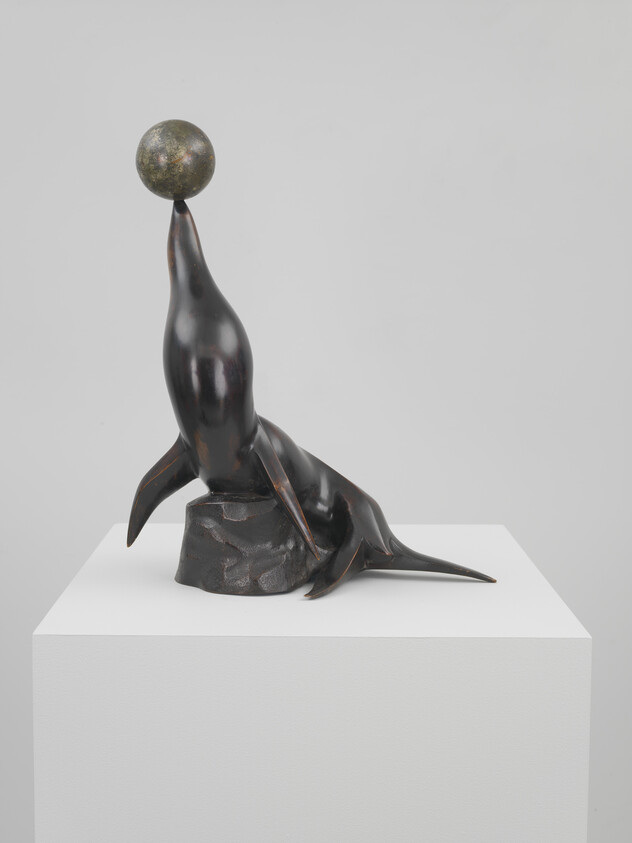 Bronze sea lion sculpture balancing a ball on its nose on a white pedestal.