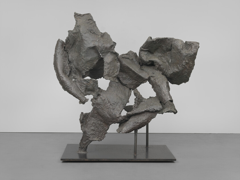 A modern abstract sculpture with a rough, textured surface in various shades of gray, displayed on a flat metal base with supporting rods, set against a plain white background in a gallery setting.