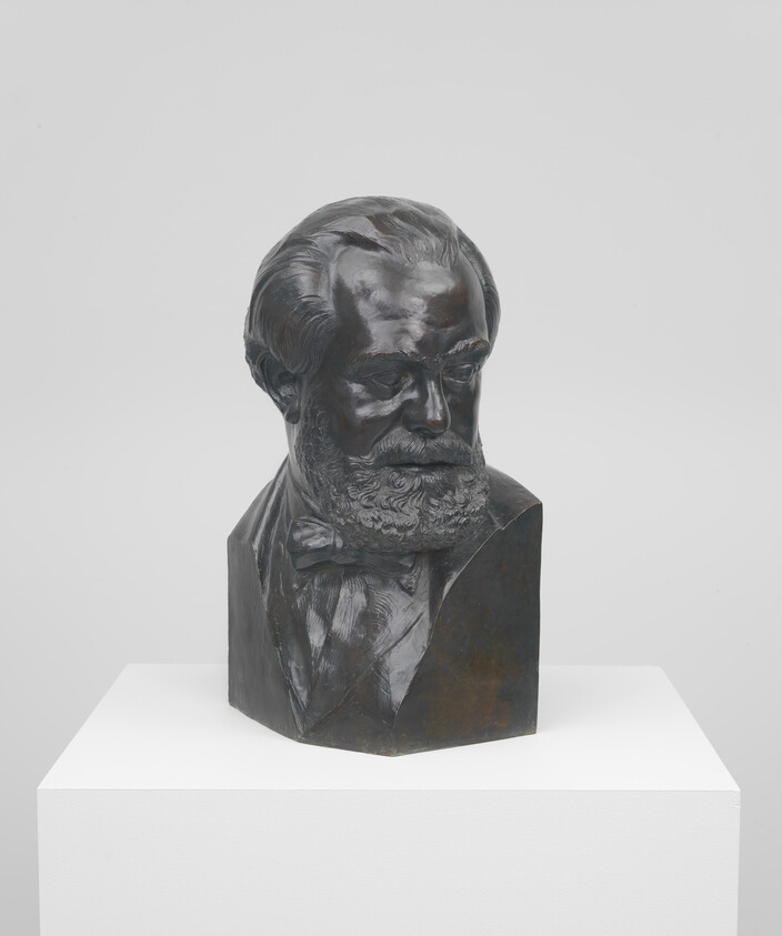 Bronze bust of a bearded man with bow tie displayed on a white pedestal.