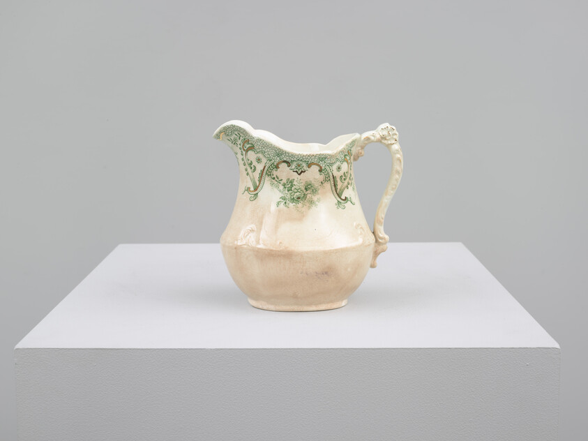 A cream-colored ceramic pitcher with green floral decoration sits on a white pedestal.