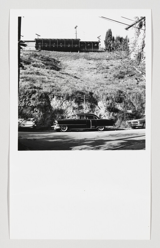 A vintage black car parked along a street in front of a grassy hillside with buildings above.