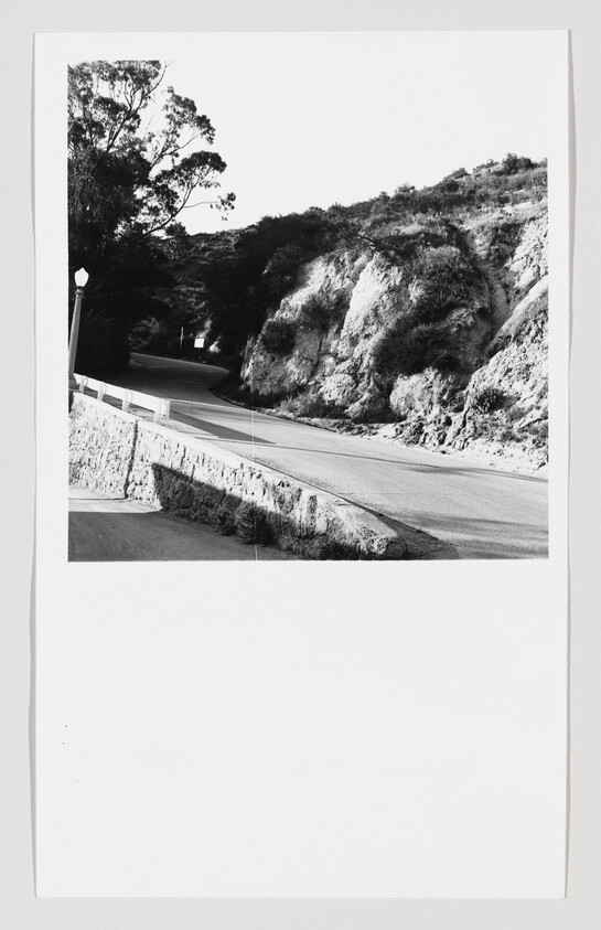 A winding road curves past rocky hills with a stone guardrail and a lamppost.