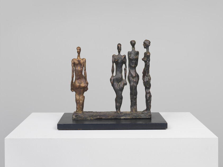 Four elongated bronze figures stand closely together on a rectangular base displayed on a pedestal.