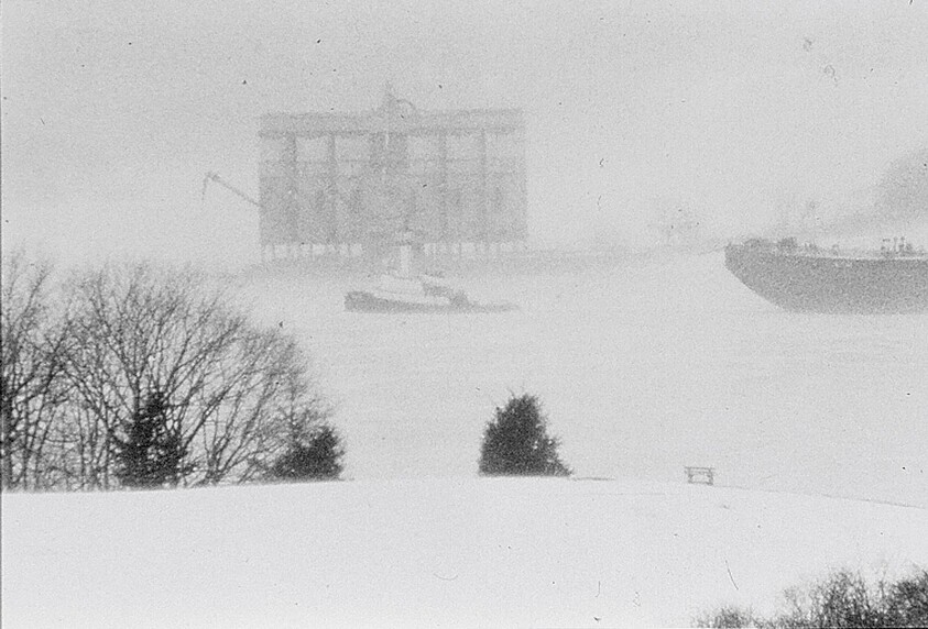Snow-covered shoreline with bench and trees overlooking a foggy river where a cargo ship and tug pass.