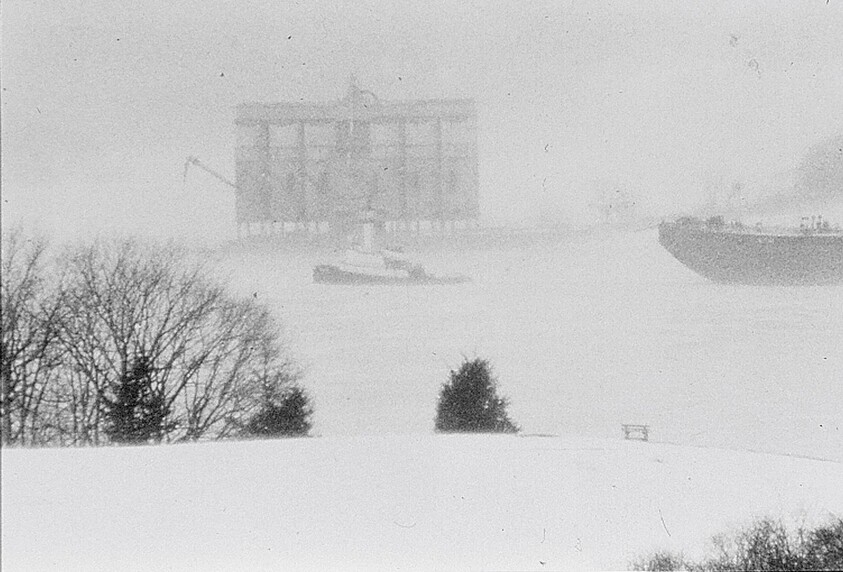 Snowy waterfront with large cargo ship and tugboat faintly visible behind trees and a bench.