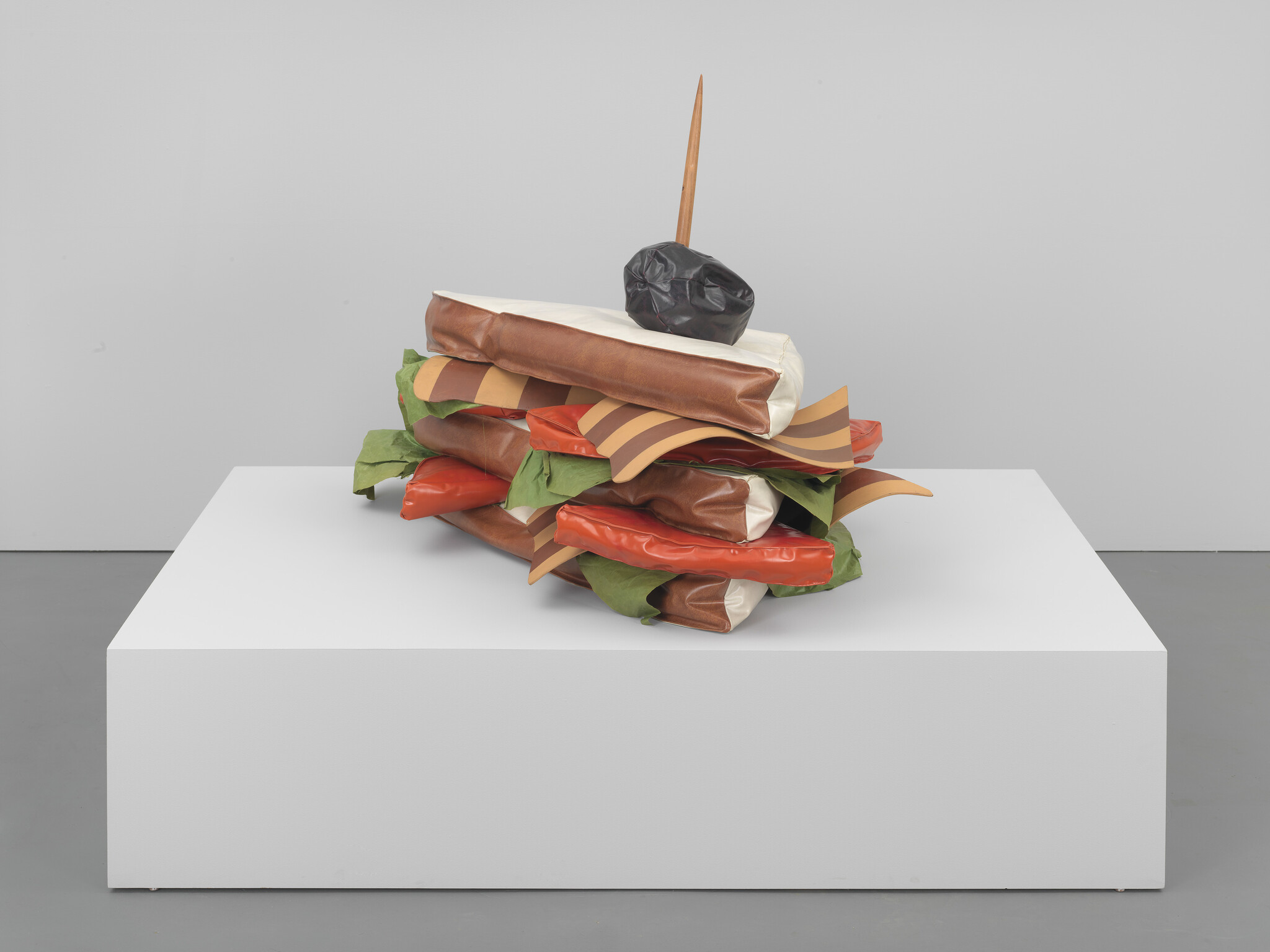 A sculpture resembling a stacked sandwich with oversized, hyperrealistic layers of lettuce, cheese, meats, and a long toothpick, displayed on a white pedestal against a grey background.