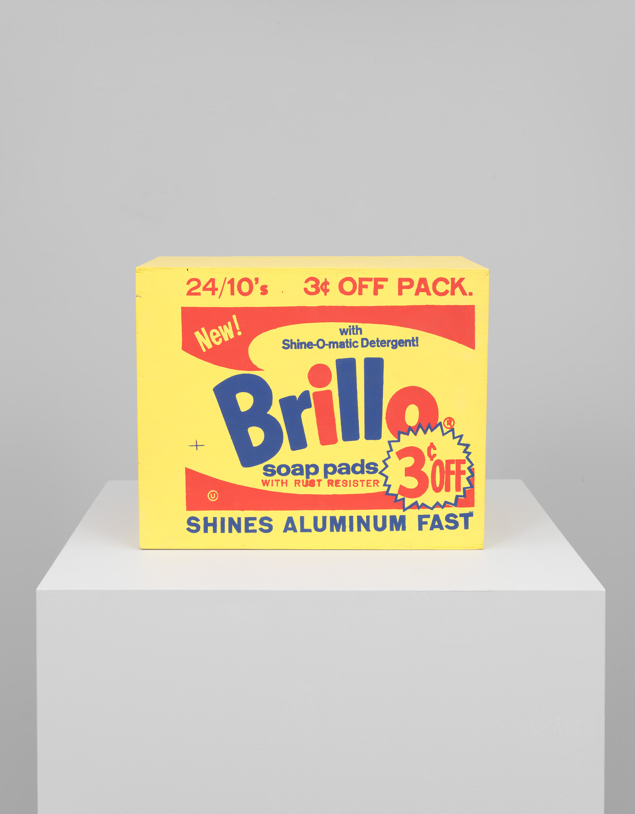 A vintage Brillo soap pads box with bold yellow and red colors advertising a 3 cent off pack, claiming to shine aluminum fast, displayed on a white pedestal against a grey background.