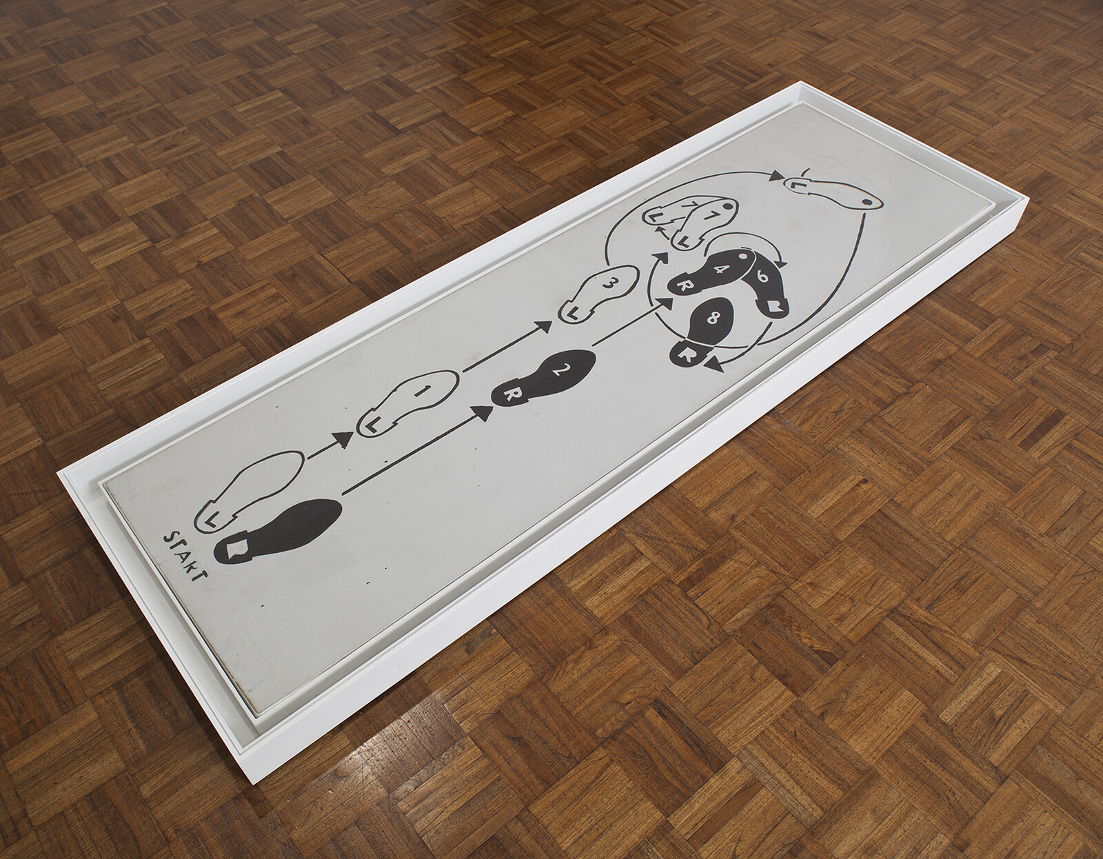 A framed piece of artwork is lying on a herringbone patterned wooden floor. The artwork features a simplistic black and white line drawing with abstract figures and shapes, including circular forms and arrows, with the word "START" at one end.