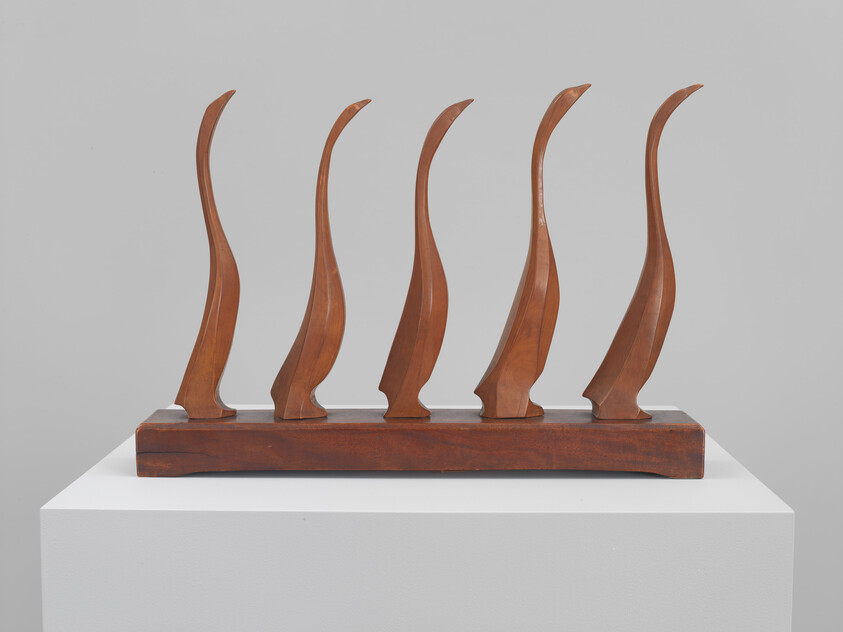 Five curved wooden flame shapes mounted on a long rectangular wooden base on a white pedestal.