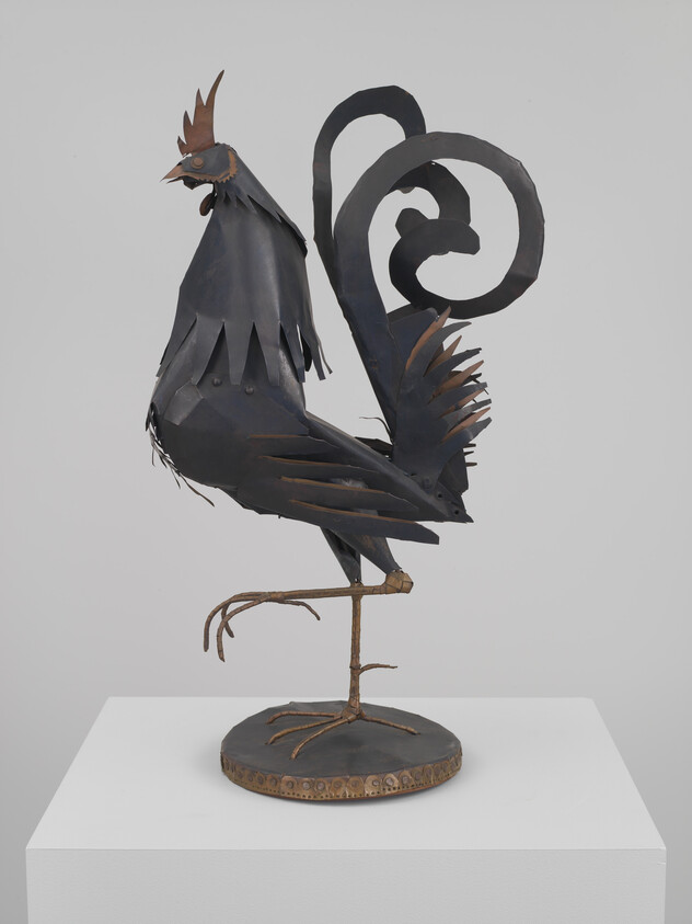 A black metal rooster sculpture stands on one leg with a curled tail on a round base.