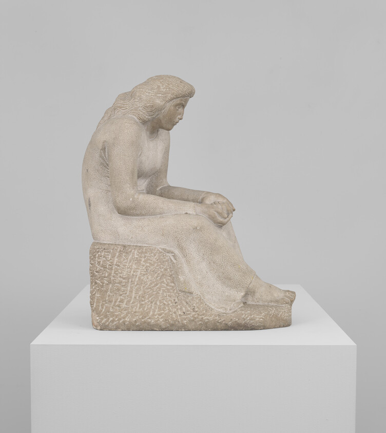 A carved stone sculpture of a seated woman looking down with hands clasped on a block.