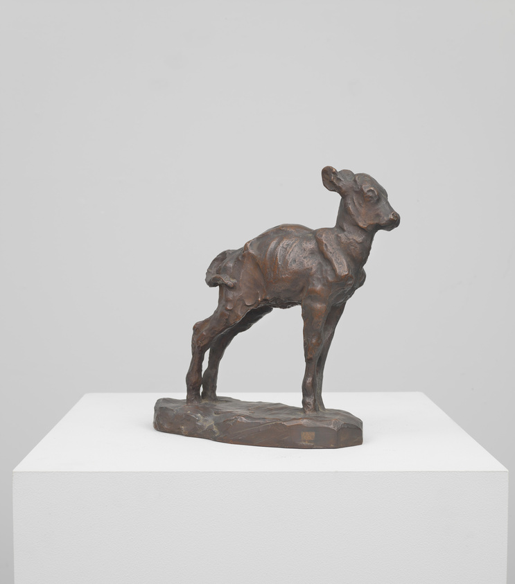 A bronze sculpture of a young deer standing on a white pedestal against a plain white background. The deer is depicted in mid-stride with one front leg lifted, suggesting movement. The sculpture shows detailed texturing to represent the animal's fur and musculature.