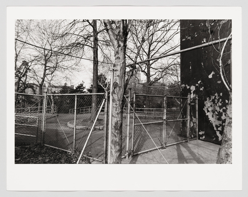 Black and white photograph depicting a fenced urban area with bare trees and a large, weathered metal structure in the background. Shadows stretch across the concrete ground, and the scene appears desolate and devoid of people.