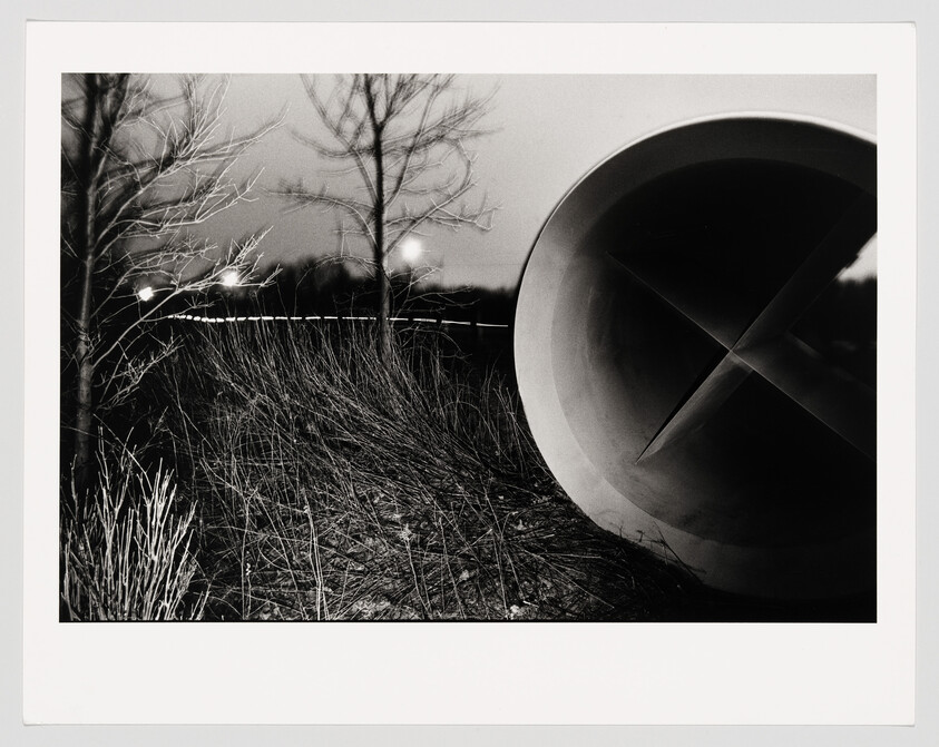 A large concrete pipe lies in a grassy field at night with bare trees and distant lights.