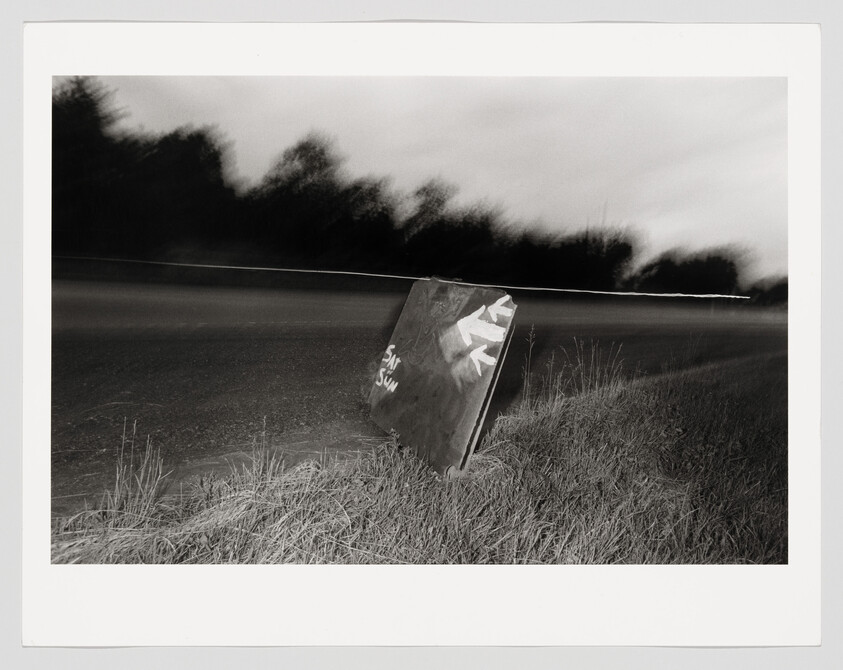 A tilted road sign with painted arrows leans beside a dark, empty roadside at night.