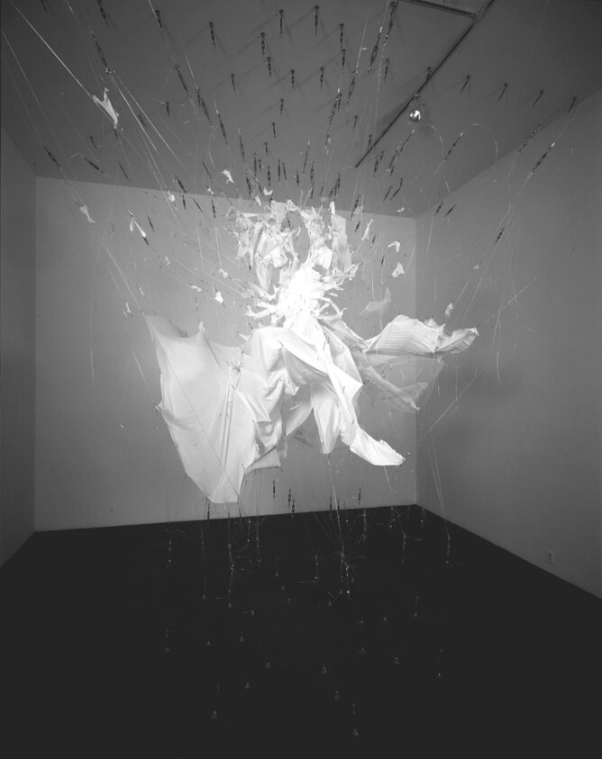 A suspended torn white fabric sculpture hangs in a dark gallery, held by many thin wires.