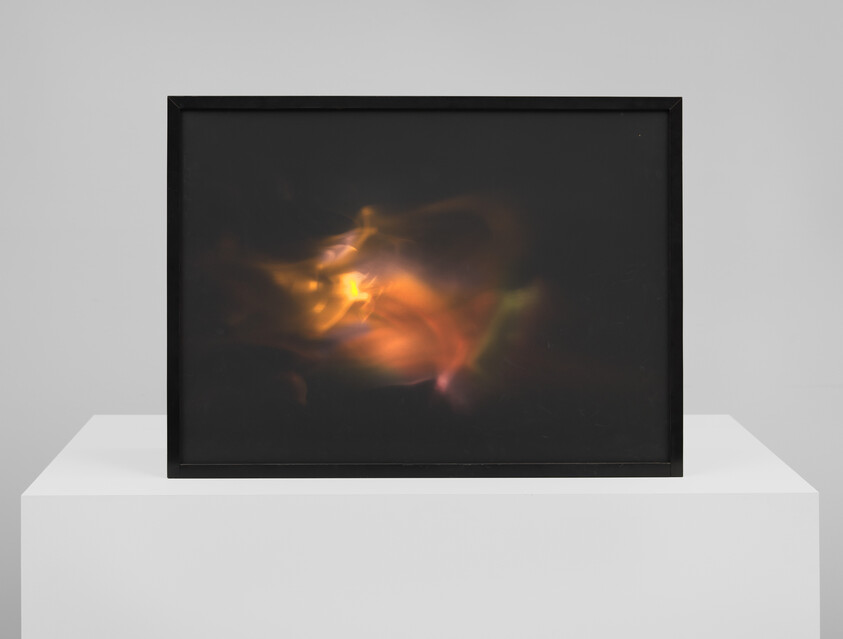 Framed dark artwork on a white pedestal showing a glowing orange and yellow abstract light form.