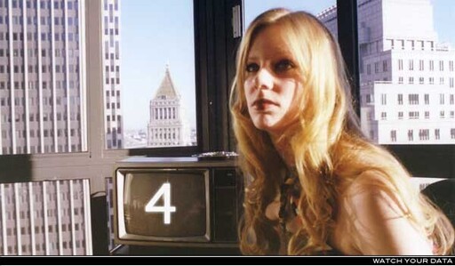 Young woman with long blonde hair sitting by a TV showing the number 4 and city skyline.