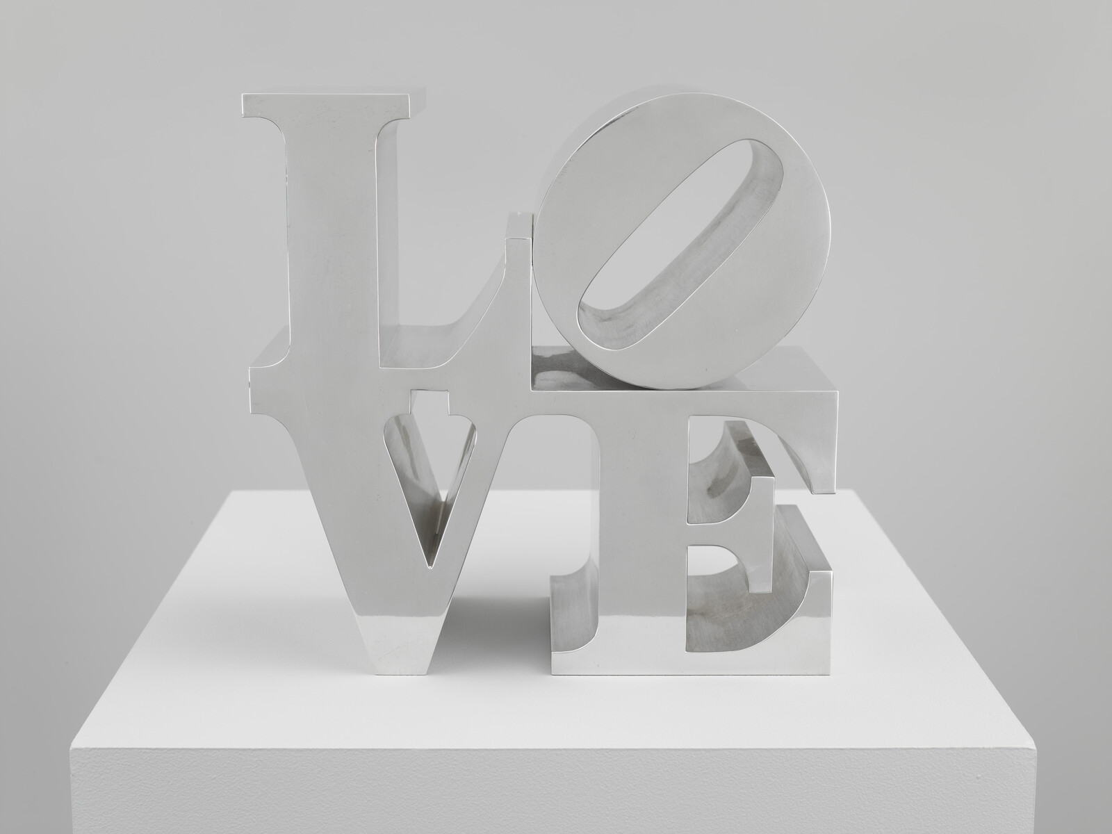 Polished metal sculpture spells the word Love with stacked letters on a white pedestal.