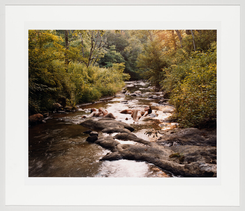 Three people relax lying on rocks in a shallow forest stream surrounded by green foliage.