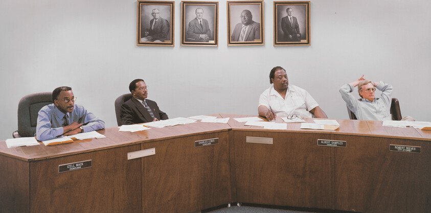 Four council members sit behind a curved wooden dais reviewing papers during a meeting.