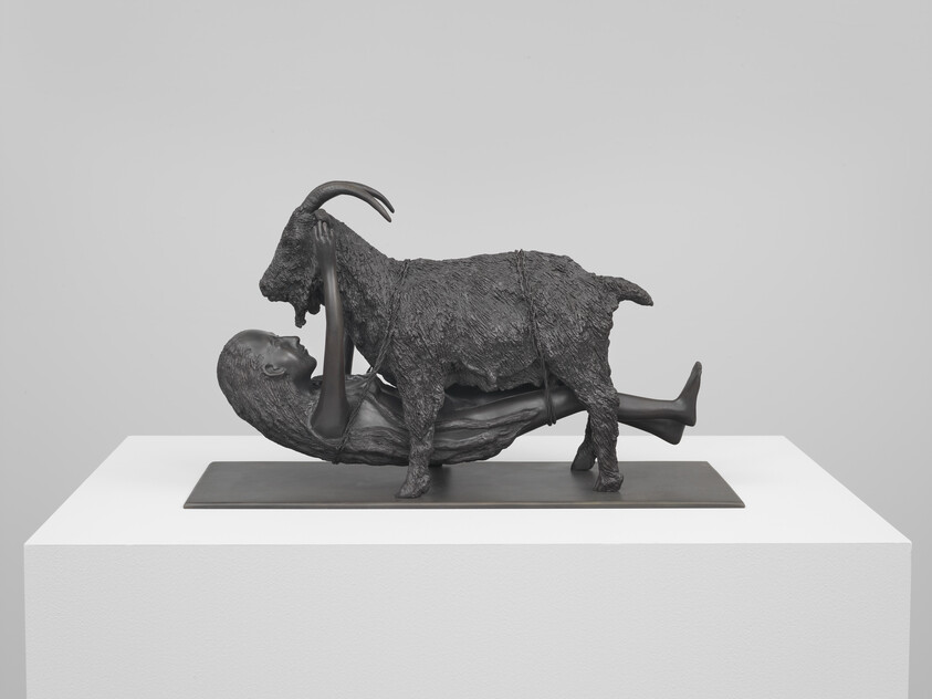 A dark bronze sculpture shows a person lying on their back supporting a goat standing on them.