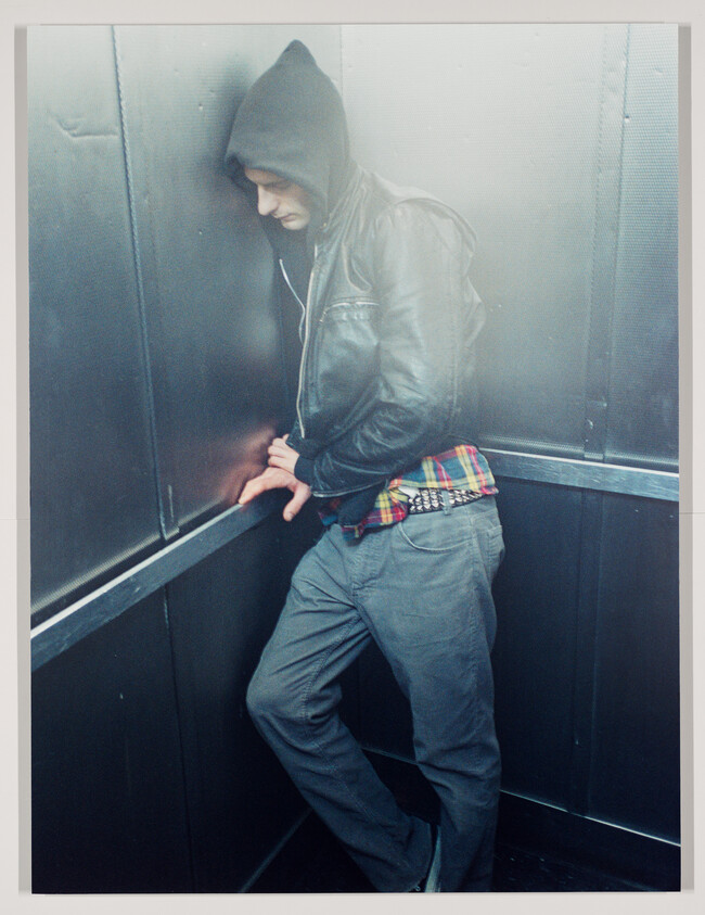 A young person in a hooded jacket leans against an elevator wall, looking down.