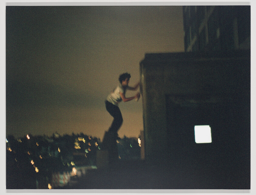 A person climbs the edge of a rooftop at night with city lights glowing below.