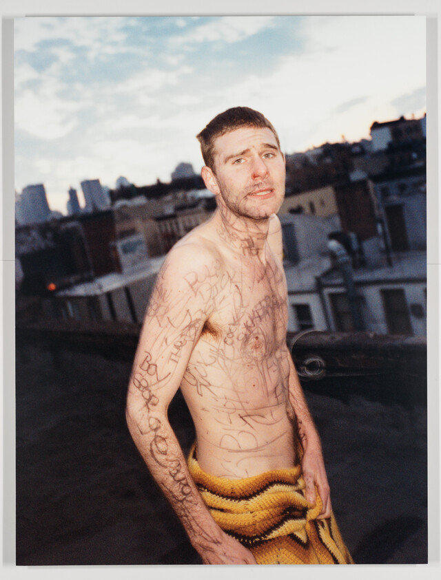 Shirtless man on a rooftop with marker doodles covering his torso, holding a yellow blanket.