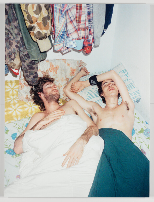 Two shirtless young men lie asleep next to each other on a bed with clothes hanging above.