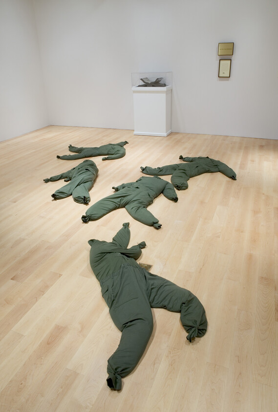 Five green, bundled fabric sculptures resembling limp human figures lie scattered across a gallery floor.