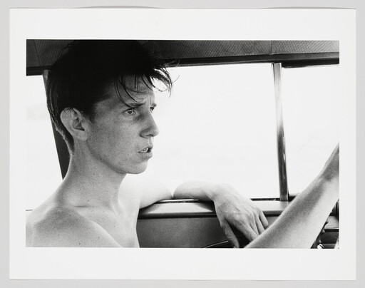 Young shirtless man driving with one arm resting on the car door and looking ahead.