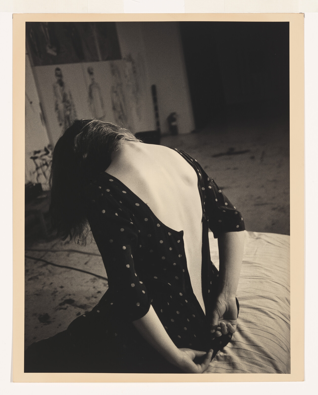 A woman with a low-back polka dot dress sits on a bed, hands clasped behind her.