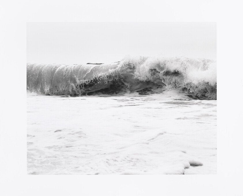 A monochrome photograph capturing a dynamic ocean wave mid-break with frothy white foam and swirling water patterns.