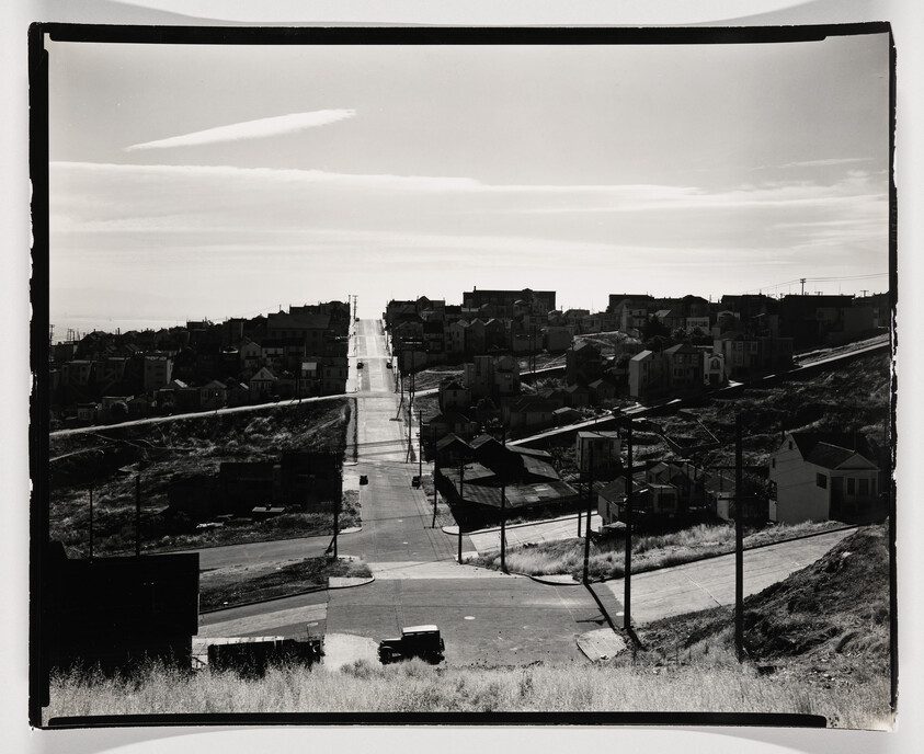 Long straight street rises through a residential hillside with houses lining both sides and a car.