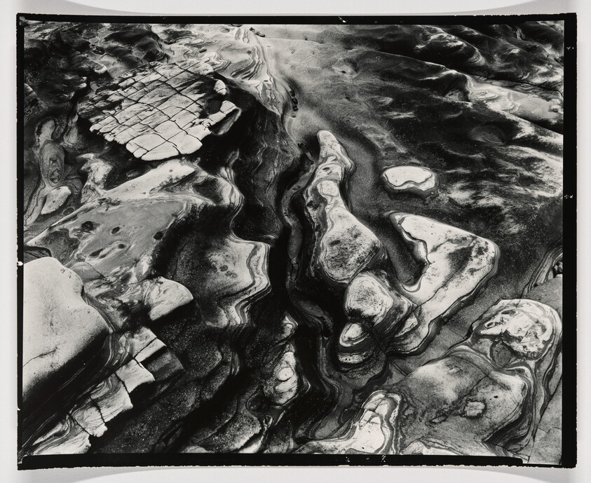 Black-and-white aerial view of smooth, swirling rock formations shaped by erosion along a shoreline.